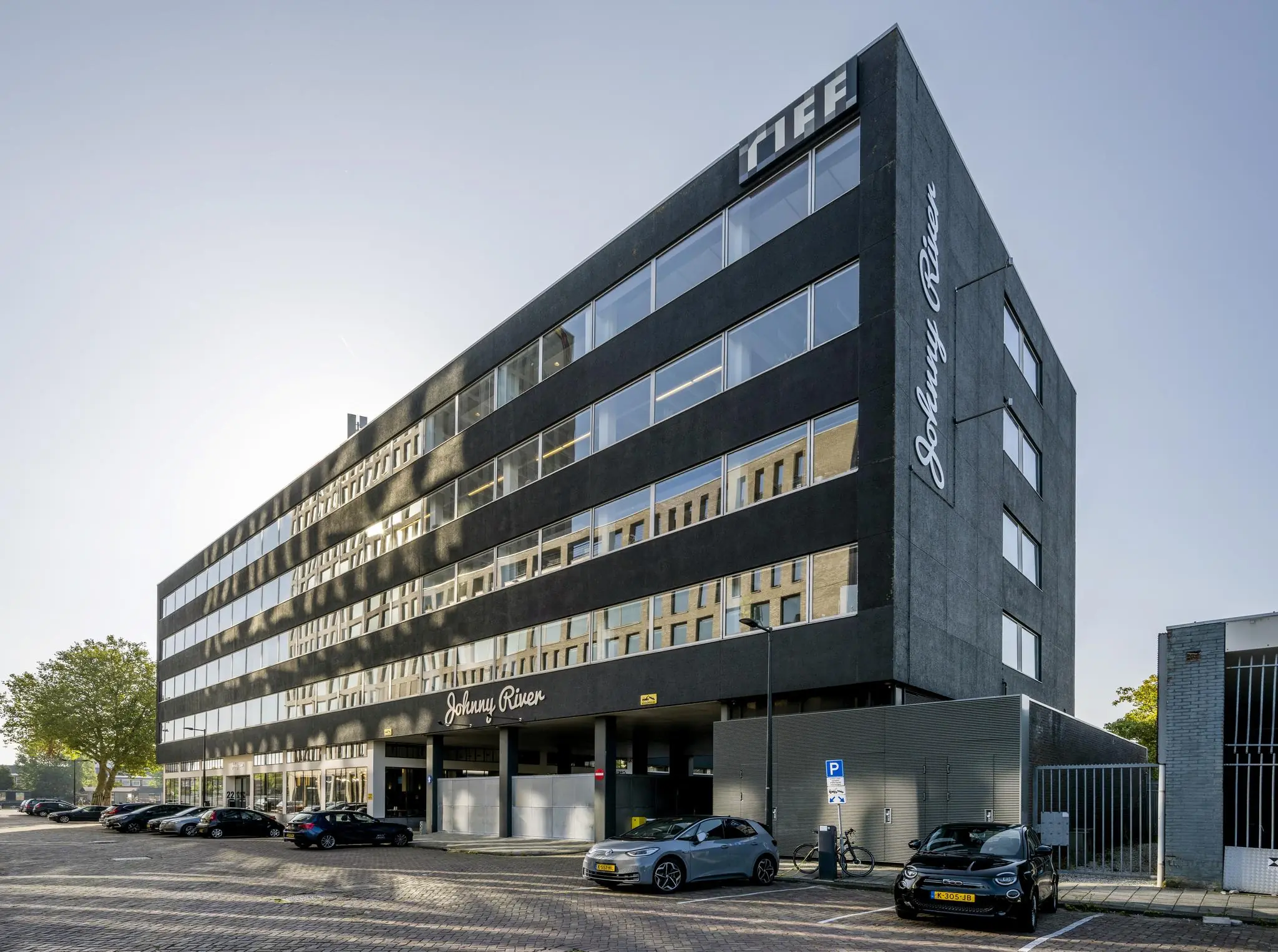 Modern zwart kantoorgebouw aan de Joan Muyskenweg met opschrift "Johnny River" en "TIFF", met geparkeerde auto’s ervoor.